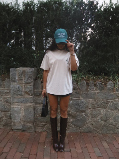 Person wearing a white oversized shirt, black shorts, and brown knee-high boots, holding a green cap.