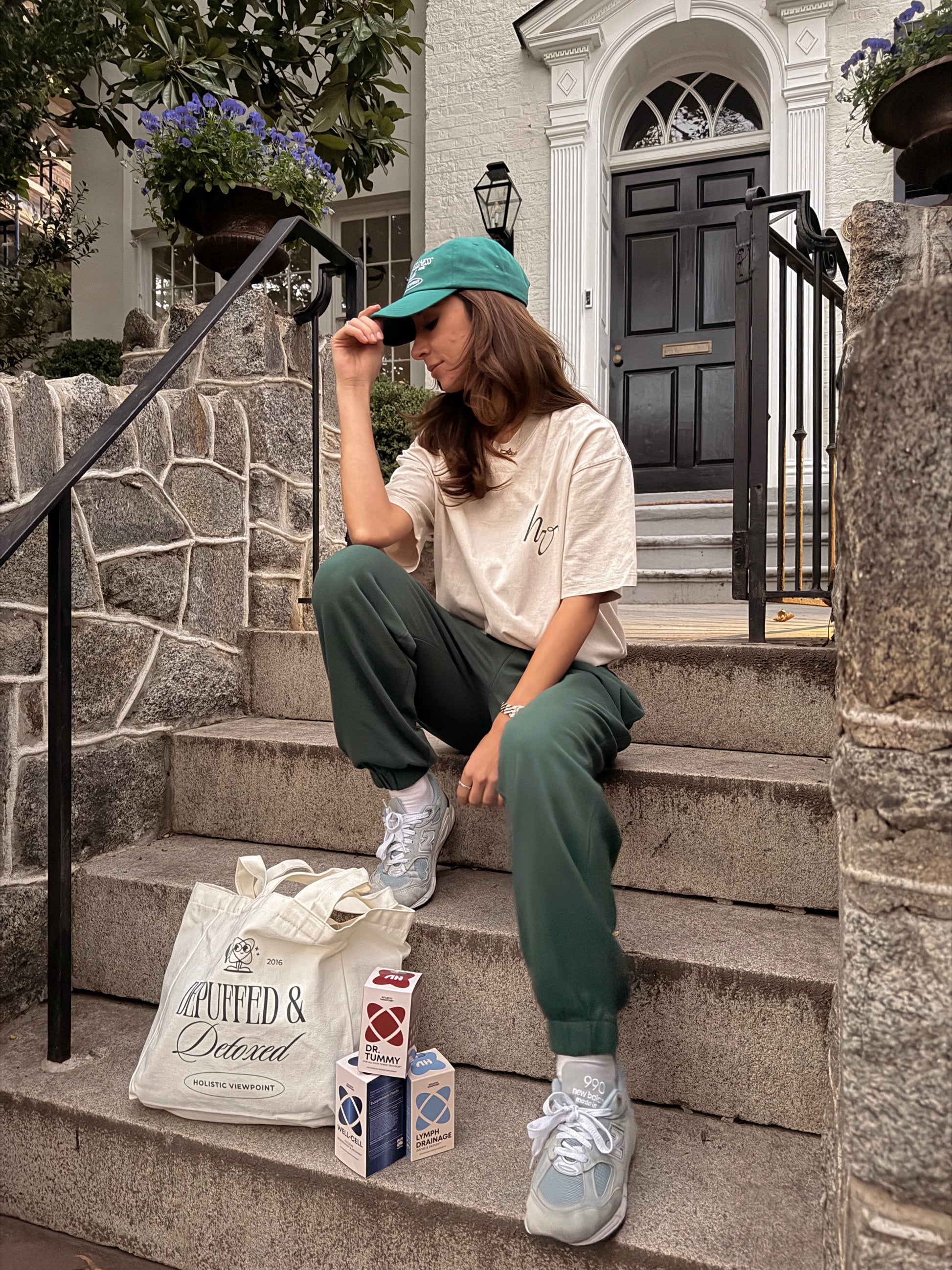 Person sitting on steps wearing a cap, white shirt, green pants, and sneakers with a bag and boxes beside them.