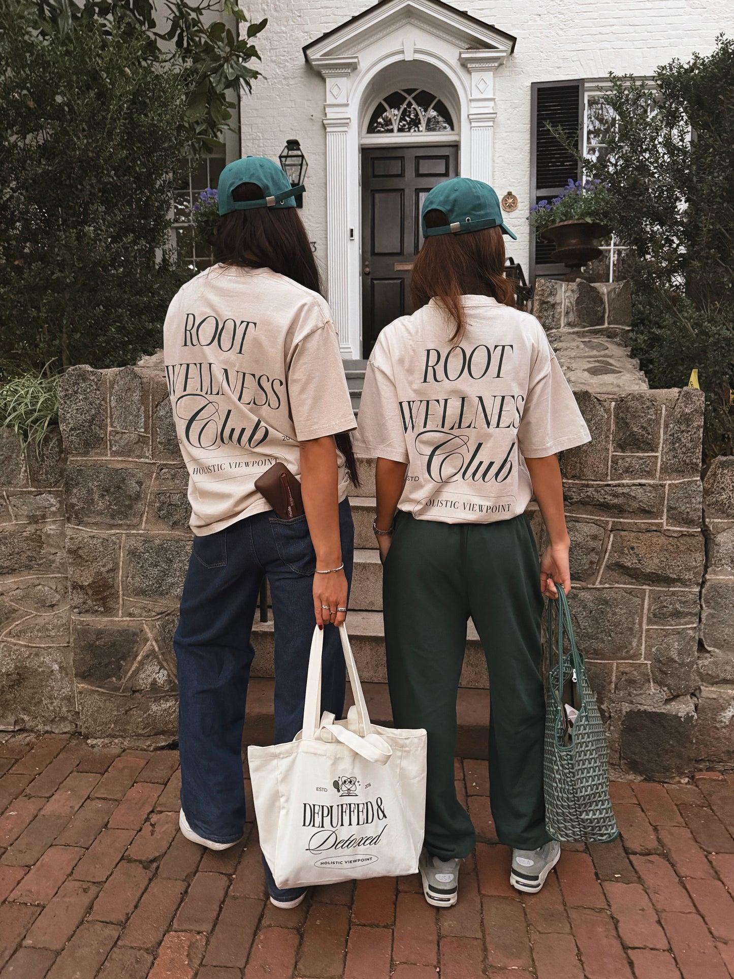 Two people wearing 'ROOT WELLNESS Club' shirts standing in front of a house.