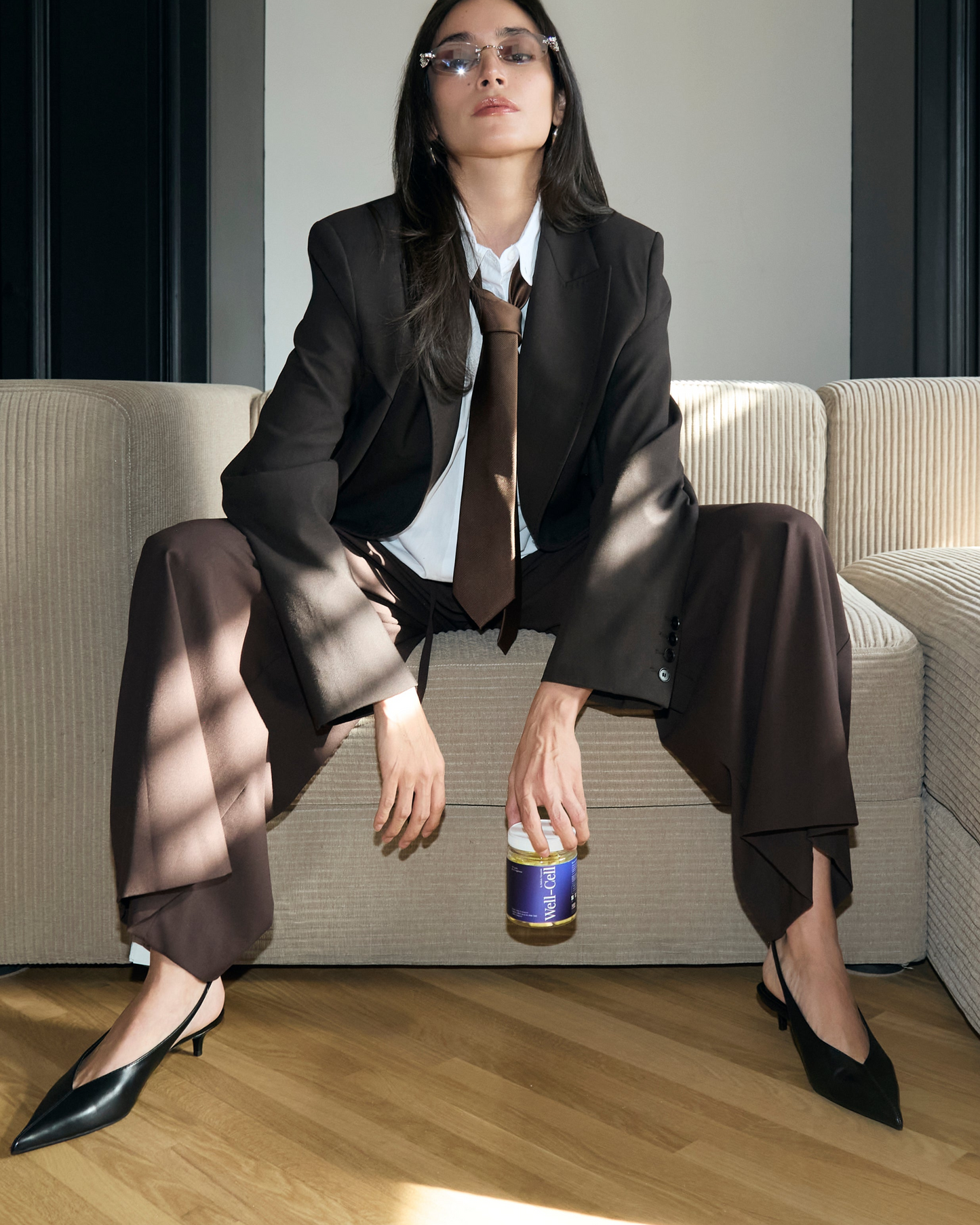 Woman in a brown suit sitting on a beige couch holding a bottle.