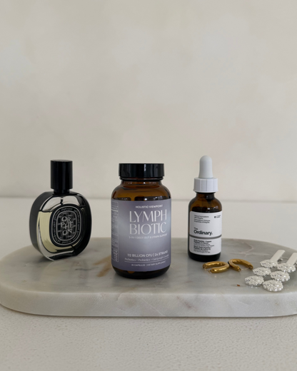 Three skincare/cosmetic products arranged on a round white marble tray against a warm off-white background: a black Diptyque bottle, a brown amber Lymphbiotic bottle, and a white dropper bottle with gold and white accessories beside it.